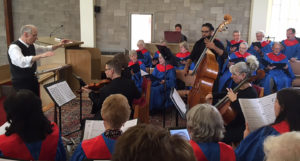 The choir, wearing red and blue robes, performing along with violin, cello, and bass players