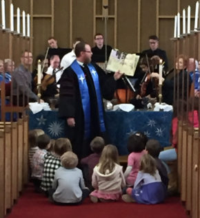 Senior Minister David Kaden holding up a book and reading to a group of youth sitting on the floor, all in front of the choir