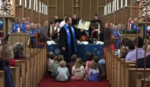 Senior Minister David Kaden holding up a book and reading to a group of youth sitting on the floor, all in front of the choir