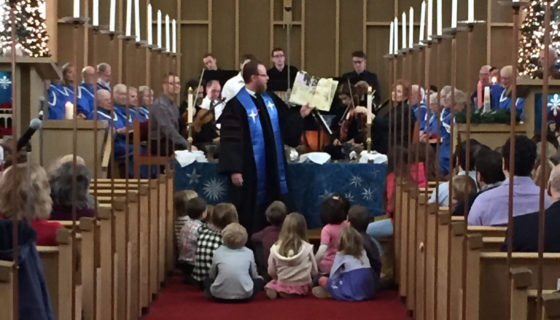 Senior Minister David Kaden holding up a book and reading to a group of youth sitting on the floor, all in front of the choir