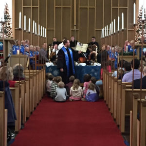 Senior Minister David Kaden holding up a book and reading to a group of youth sitting on the floor, all in front of the choir
