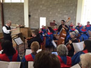 The choir, wearing red and blue robes, performing along with violin, cello, and bass players