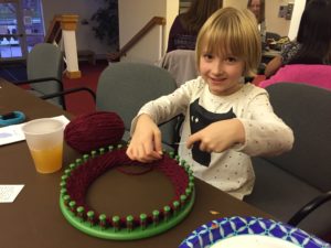 A girl knitting with maroon yarn on a loom