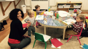 Three toddlers standing around a table painting in the nursery along with an adult volunteer
