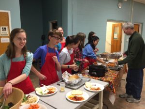 A group of youth wearing red aprons serving the meal at a pasta dinner fundraiser in the fellowship hall