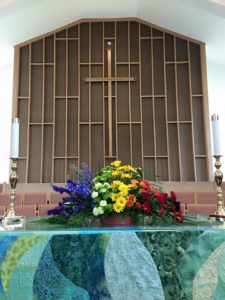 A rainbow-colored flower arrangement on the altar in front of the cross on the sanctuary wall