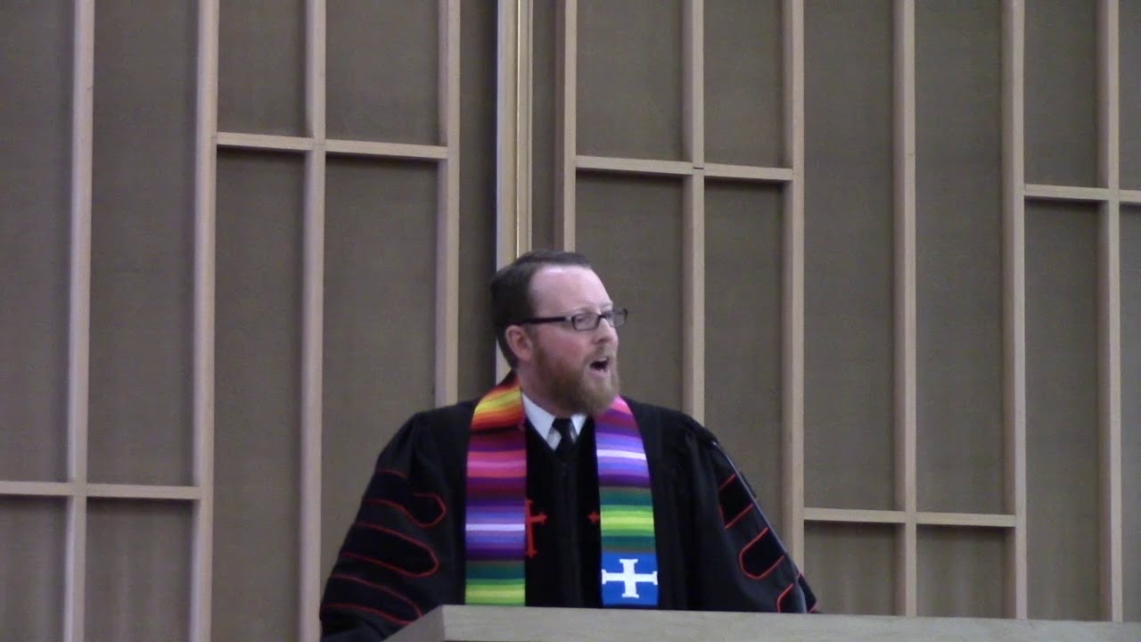 Senior Minister David Kaden preaching from the pulpit and wearing a black robe and multicolored stole
