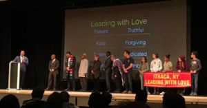 13 community leaders, faith leaders, and students (including Mayor Myrick, Superintendent Brown, and Pastor Kaden) on a stage with a banner reading "Ithaca, NY Leading with Love"